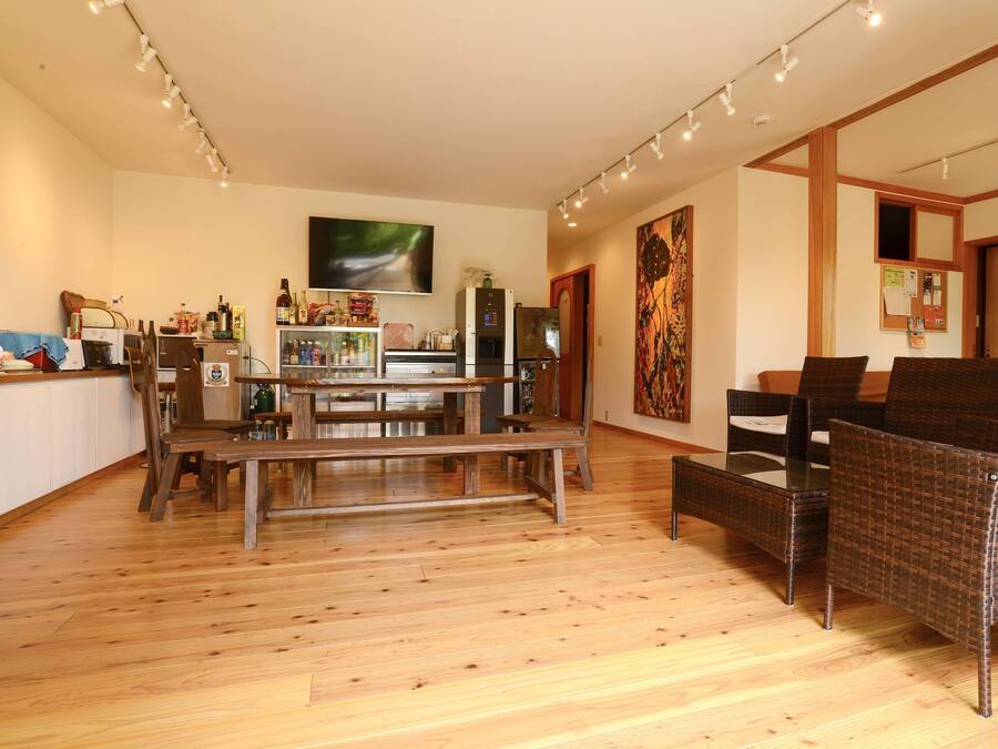 A spacious living area with a wooden floor, a dining table, wicker furniture, and a kitchen area with appliances and a refrigerator.