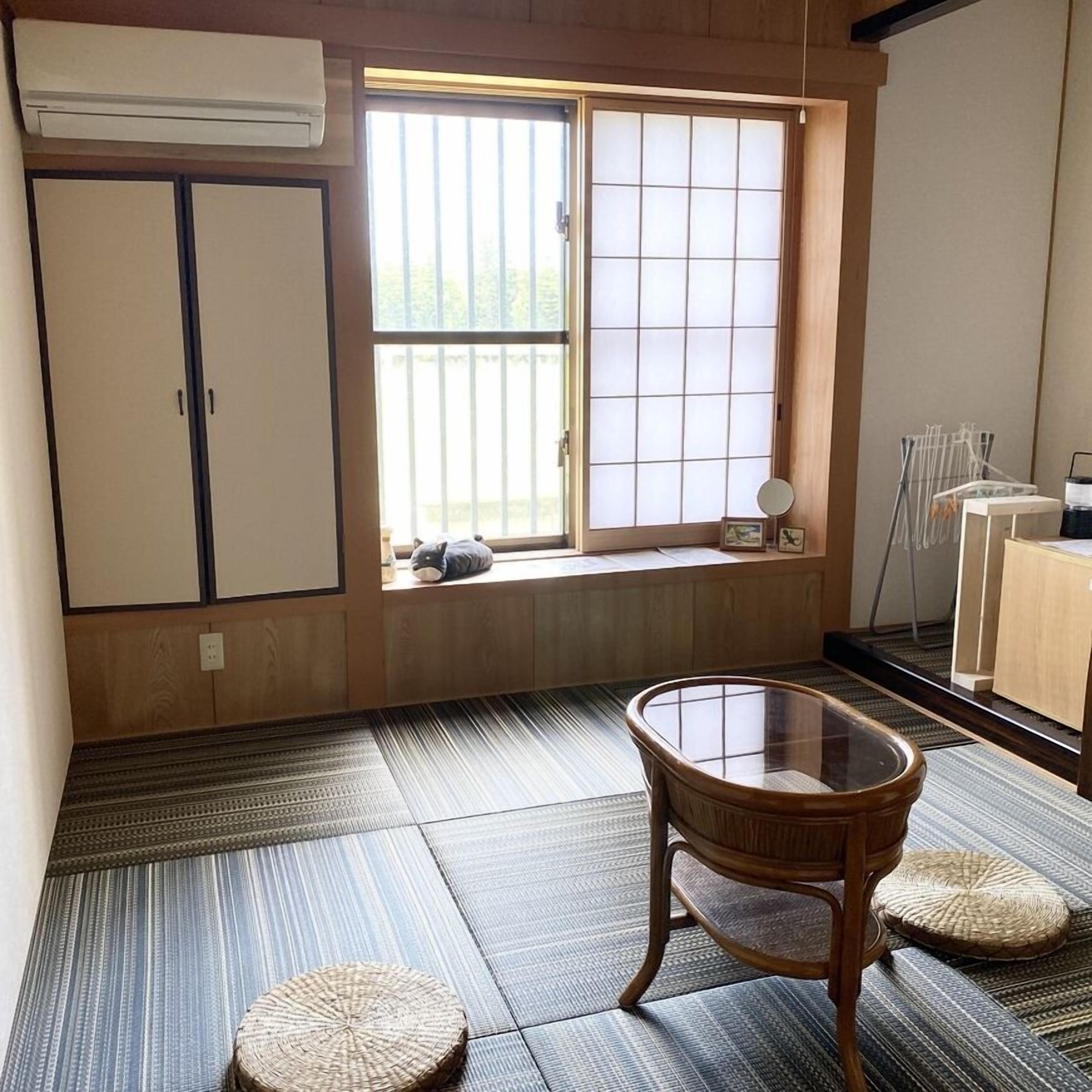 A room with tatami flooring, a sliding door, a small table with a glass top, and a window with a view of greenery.