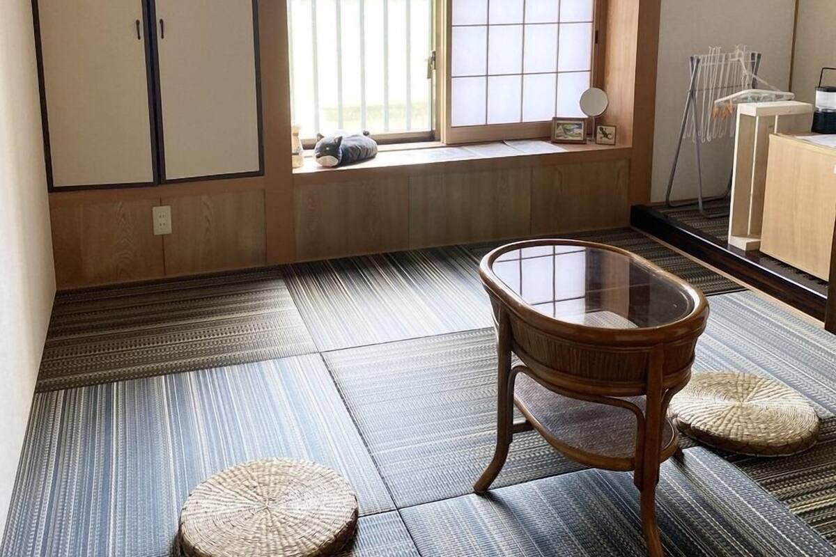 A room with tatami flooring, a sliding door, a small table with a glass top, and a window with a view of greenery.
