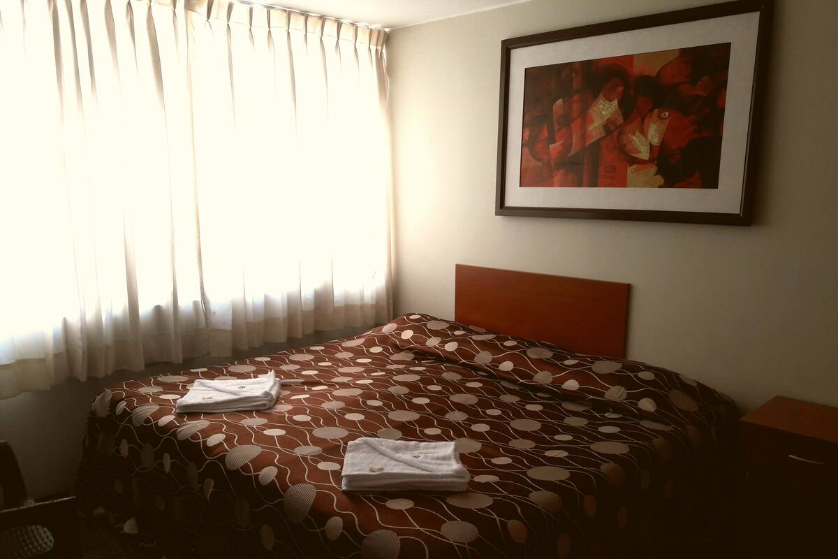 Ein ordentlich bezogenes Bett mit einem gemusterten Überwurf, ein gerahmtes Kunstwerk an der Wand und ein Fenster mit durchsichtigen Vorhängen.