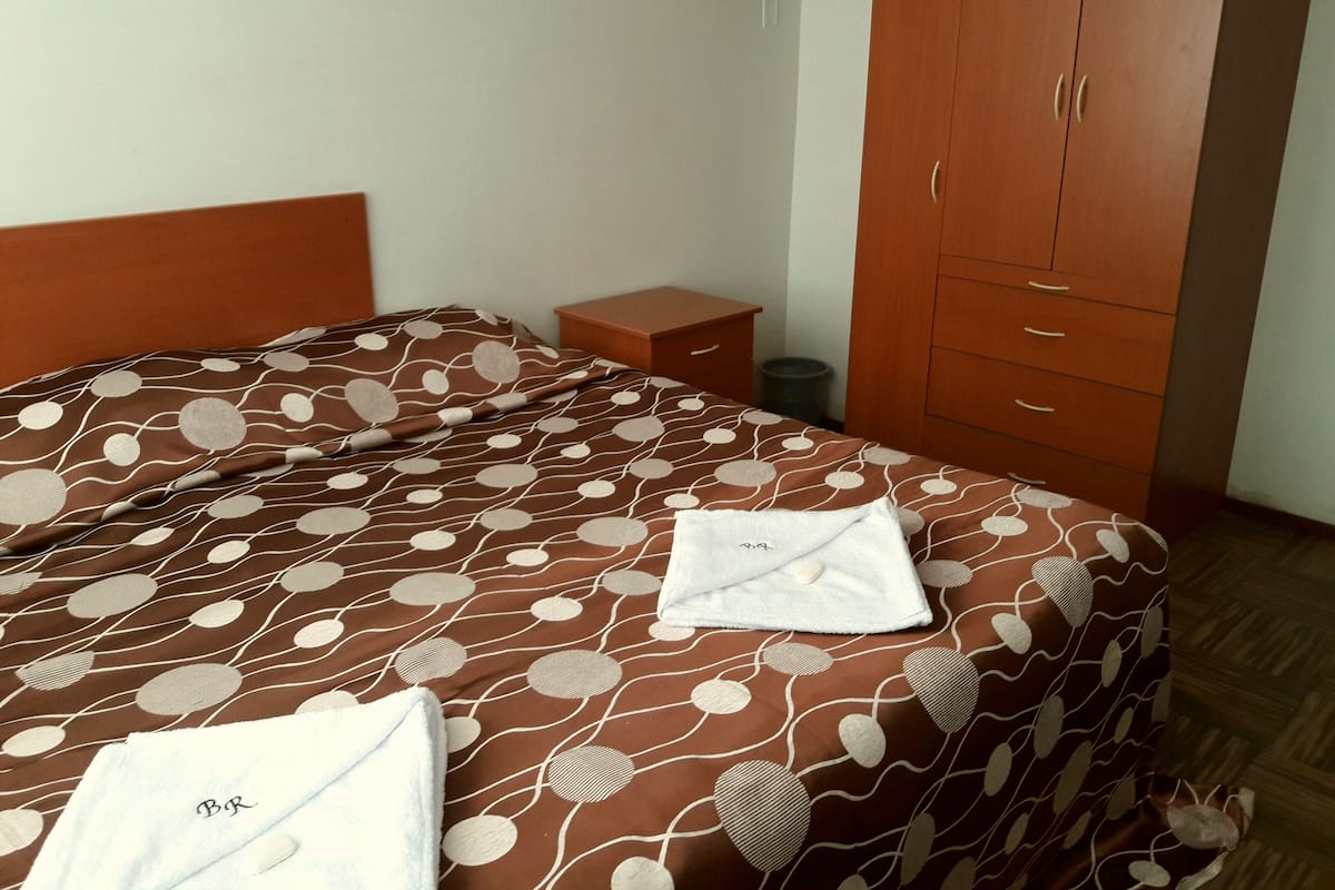Ein ordentlich bezogenes Bett mit einem gemusterten Überwurf, ein kleiner Nachttisch, ein Kleiderschrank und ein Mülleimer.