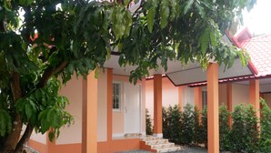 Un edificio con pareti arancioni, tetto rosso e terreno sterrato. Scale conducono a una porta, circondata da vegetazione.
