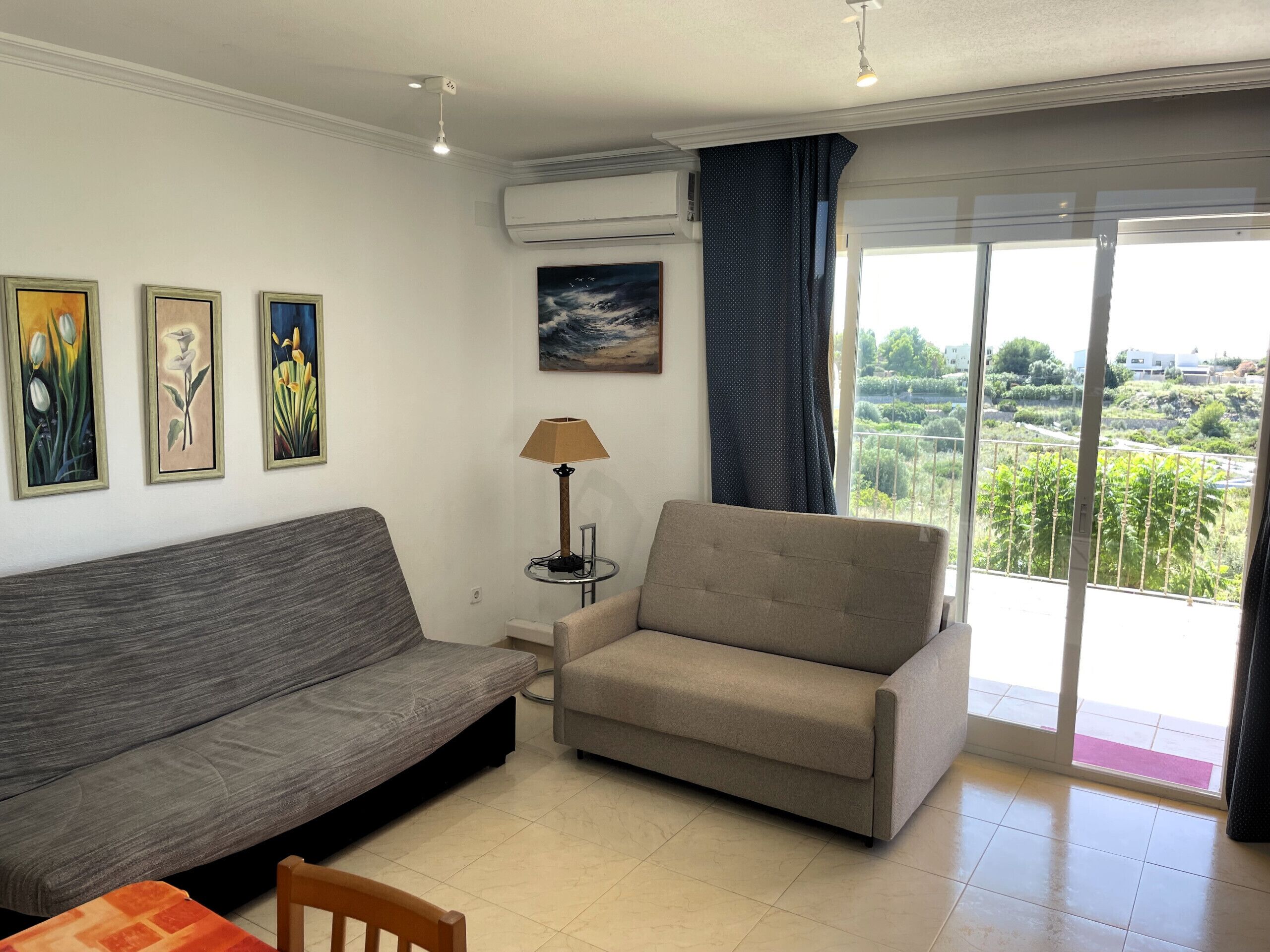 Un salon avec un canapé gris, un fauteuil beige et une petite table sur laquelle se trouve une lampe. Trois œuvres d’art encadrées ornent le mur, et une grande fenêtre offre une vue sur la verdure et les bâtiments.