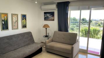 Un salon avec un canapé gris, un fauteuil beige et une petite table sur laquelle se trouve une lampe. Trois œuvres d’art encadrées ornent le mur, et une grande fenêtre offre une vue sur la verdure et les bâtiments.