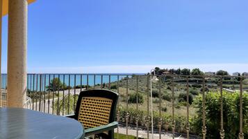 Een balkon met een tafel en stoel, met uitzicht op een kustlandschap en een helder zicht op de zee en verre gebouwen.