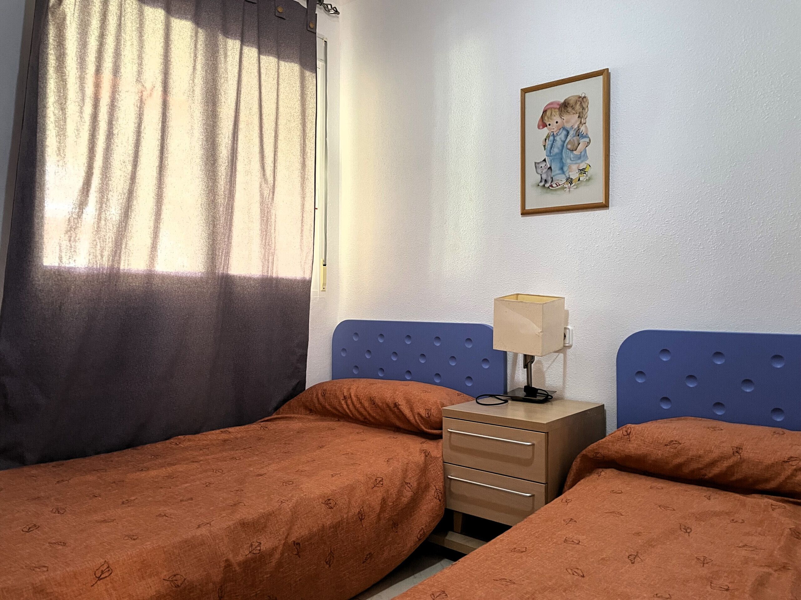 Una habitación con dos camas, una mesita de noche con lámpara, un cuadro en la pared y una ventana con cortinas.