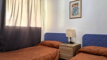 Una habitación con dos camas, una mesita de noche con lámpara, un cuadro en la pared y una ventana con cortinas.
