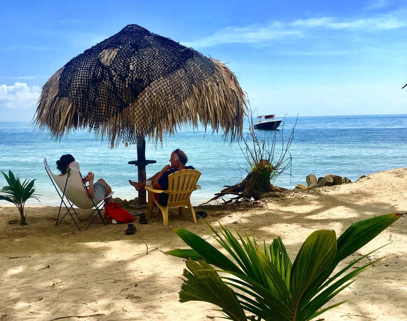 Escena de playa con una sombrilla de paja, dos sillas y una mesa pequeña.