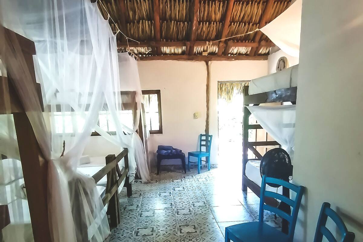 Une pièce avec un toit de chaume, un lit doté d’une moustiquaire, une chaise bleue et des carreaux de sol à motifs.