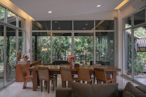 A spacious dining area with a wooden table and chairs, large windows offering a view of greenery outside.