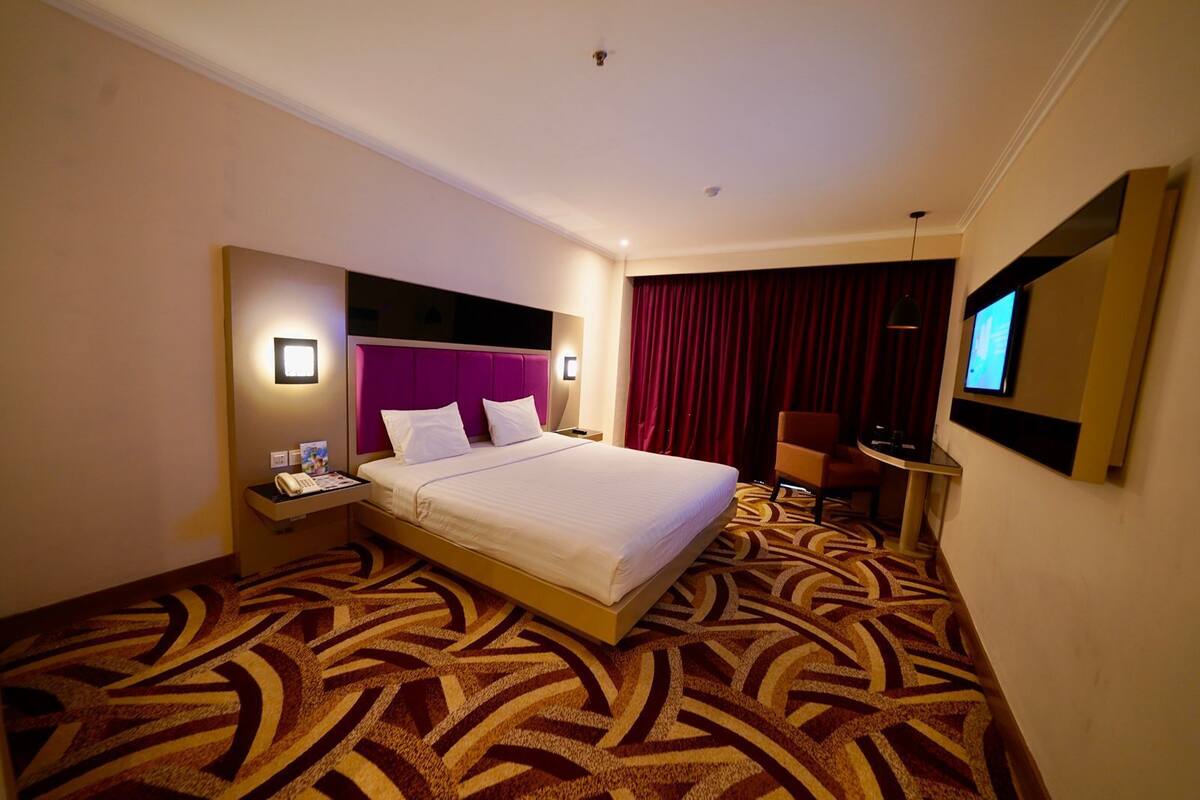 A hotel room with a large bed, a nightstand with a phone and magazine, a chair, a TV, and a patterned carpet.