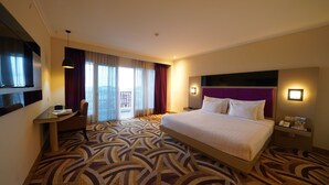 A hotel room with a large bed, a desk, a chair, a TV, and a balcony with curtains. - Ayola Sentosa Palembang (Palembang)