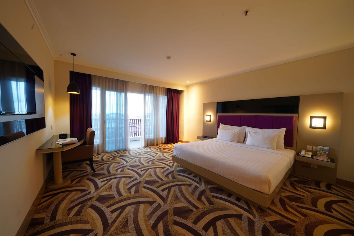 A hotel room with a large bed, a desk, a chair, a TV, and a balcony with curtains.