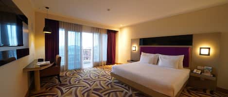 A hotel room with a large bed, a desk, a chair, a TV, and a balcony with curtains.