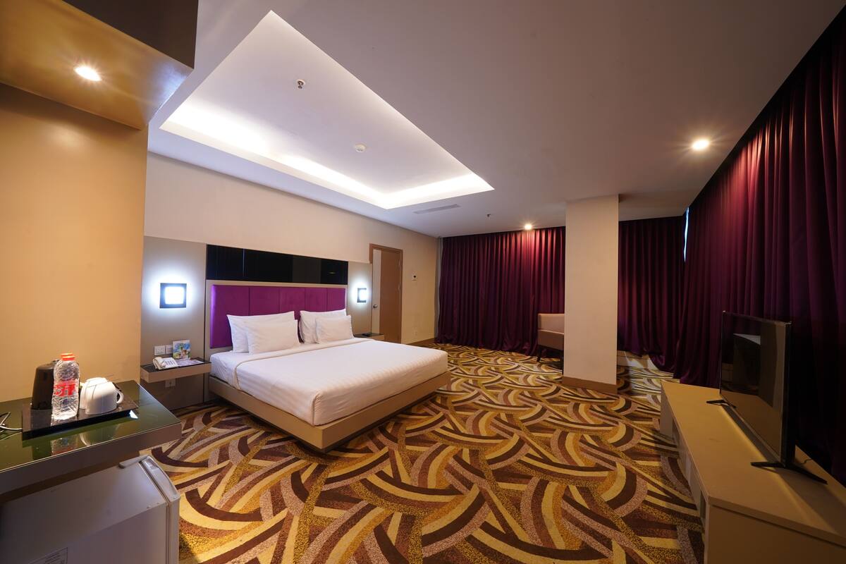 A hotel room with a bed, a desk with a computer, a TV, a minibar, and a bathroom.