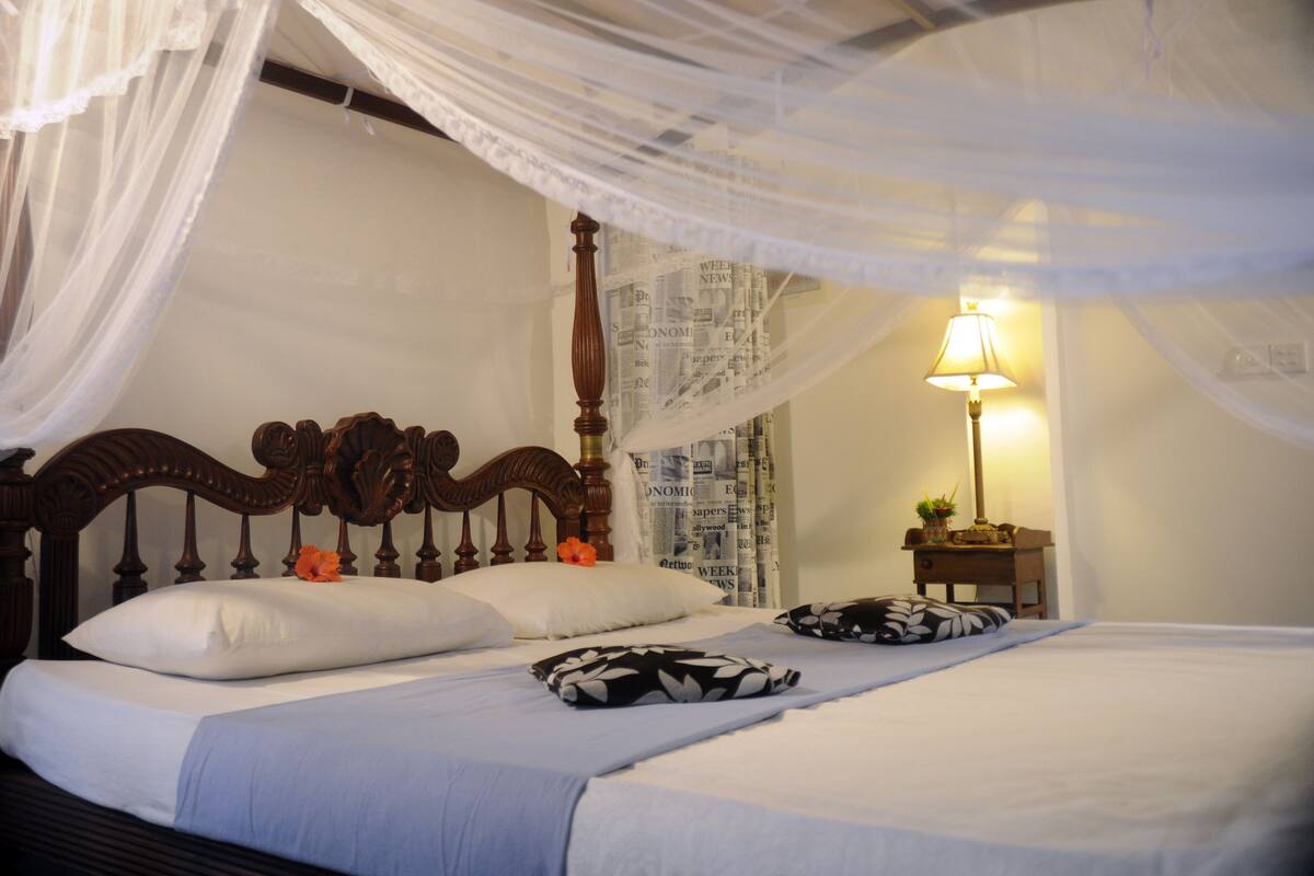 A bed with a canopy, a bedside table with a lamp, and a wall with newspaper wallpaper.