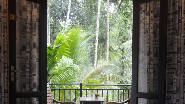 A room with a glass door leading to a balcony with a view of a forest. There is a small table and two chairs on the balcony. - Banja Little Villa (Hikkaduwa)