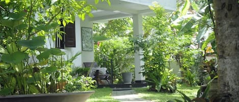 A garden pathway leading to a covered area with a bench and a table, surrounded by lush greenery and a white building.
