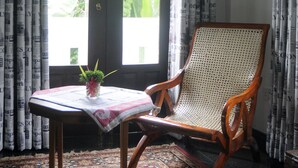 A room with a wooden chair, a small table with a plant, and a patterned rug. There is a window with curtains and a door with a decorative design. - Banja Little Villa (Hikkaduwa)