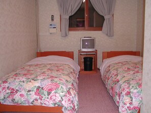 Une petite chambre agréable, dotée de deux lits simples, d’une télévision posée sur une petite table et d’une fenêtre avec des rideaux.