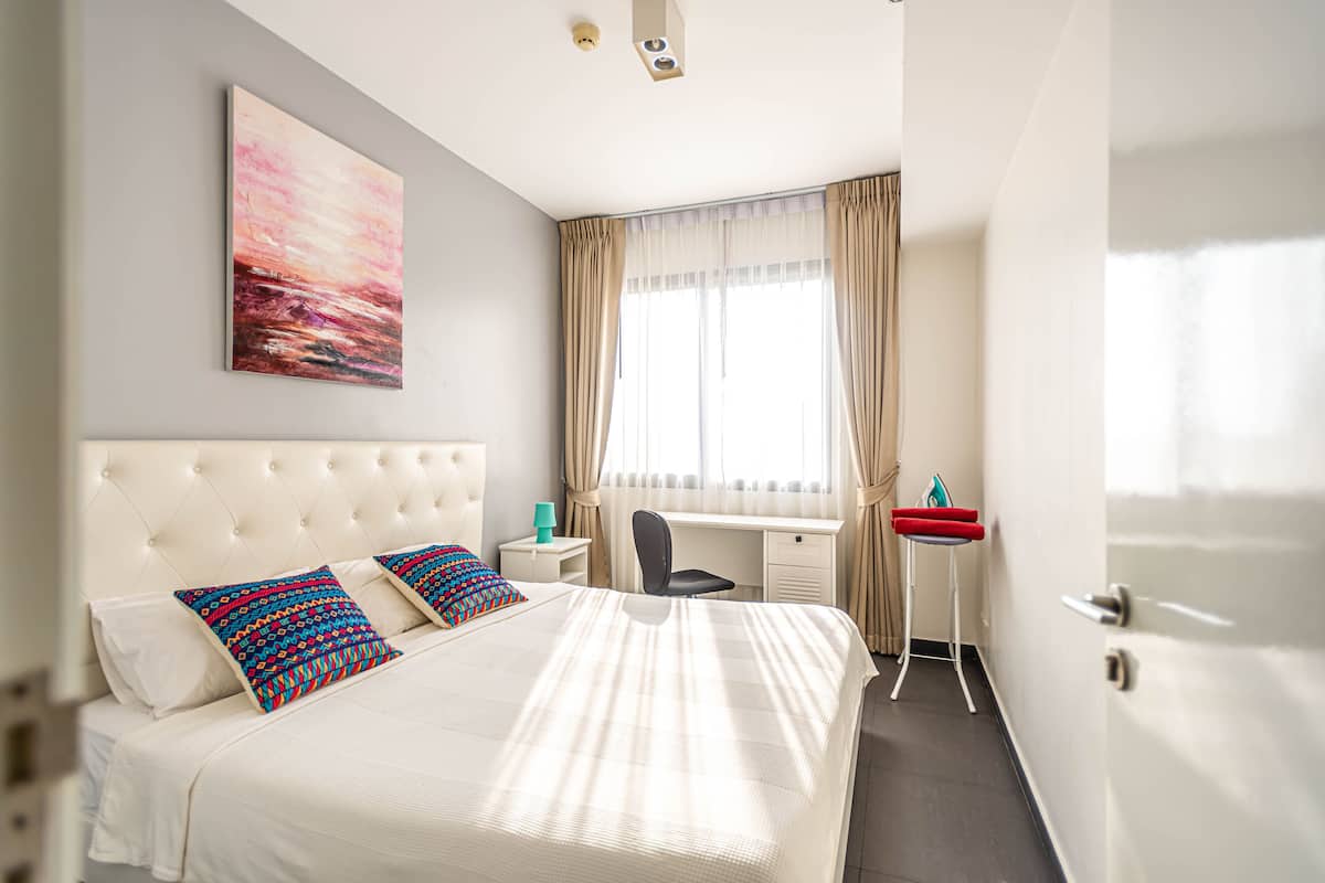 A hotel room with a large bed, a desk, and a chair. There is a window with curtains and a painting on the wall.