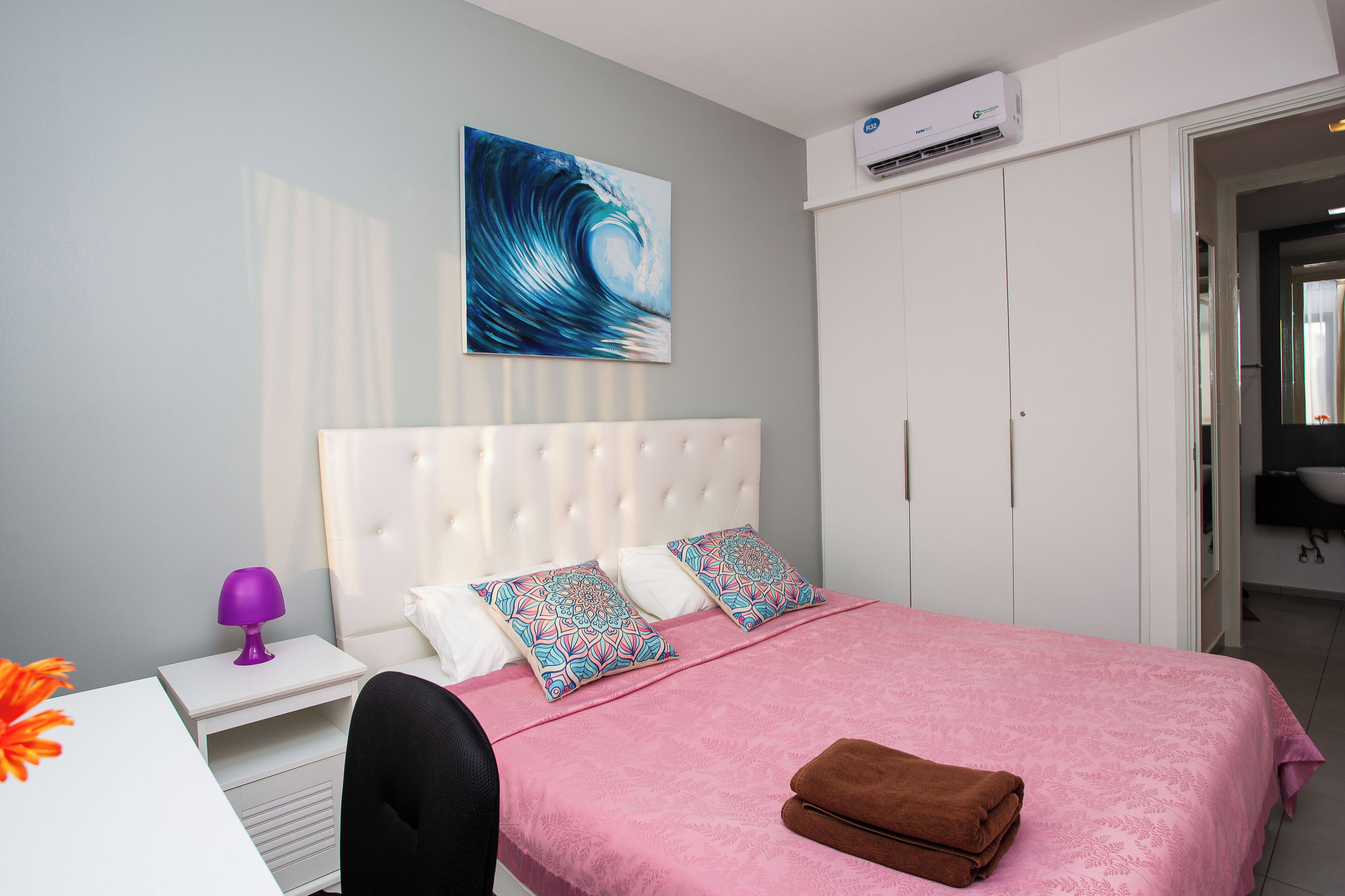 A bedroom with a bed, white headboard, two patterned pillows, a pink bedspread, a brown towel, a nightstand with a purple lamp, and a wall-mounted air conditioner.