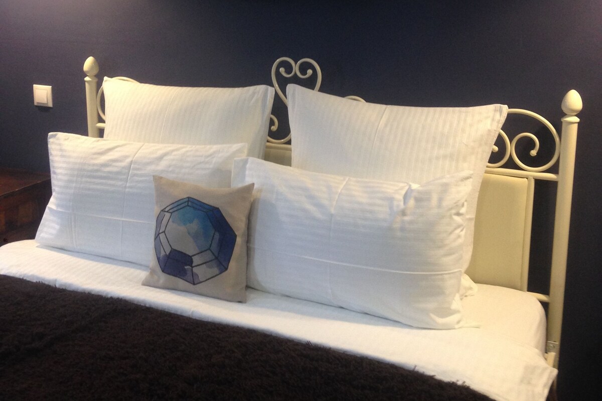 Una cama con ropa de cama blanca y una almohada geométrica distintiva, frente a una pared oscura con un cuadro enmarcado arriba.