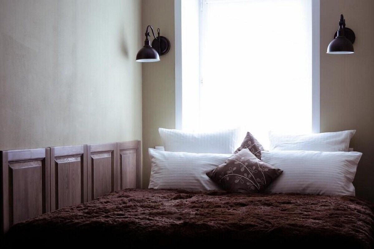 Un dormitorio con cabecera de madera, un ventanal grande con dos lámparas de pared, y una cama con almohadas blancas y estampadas.