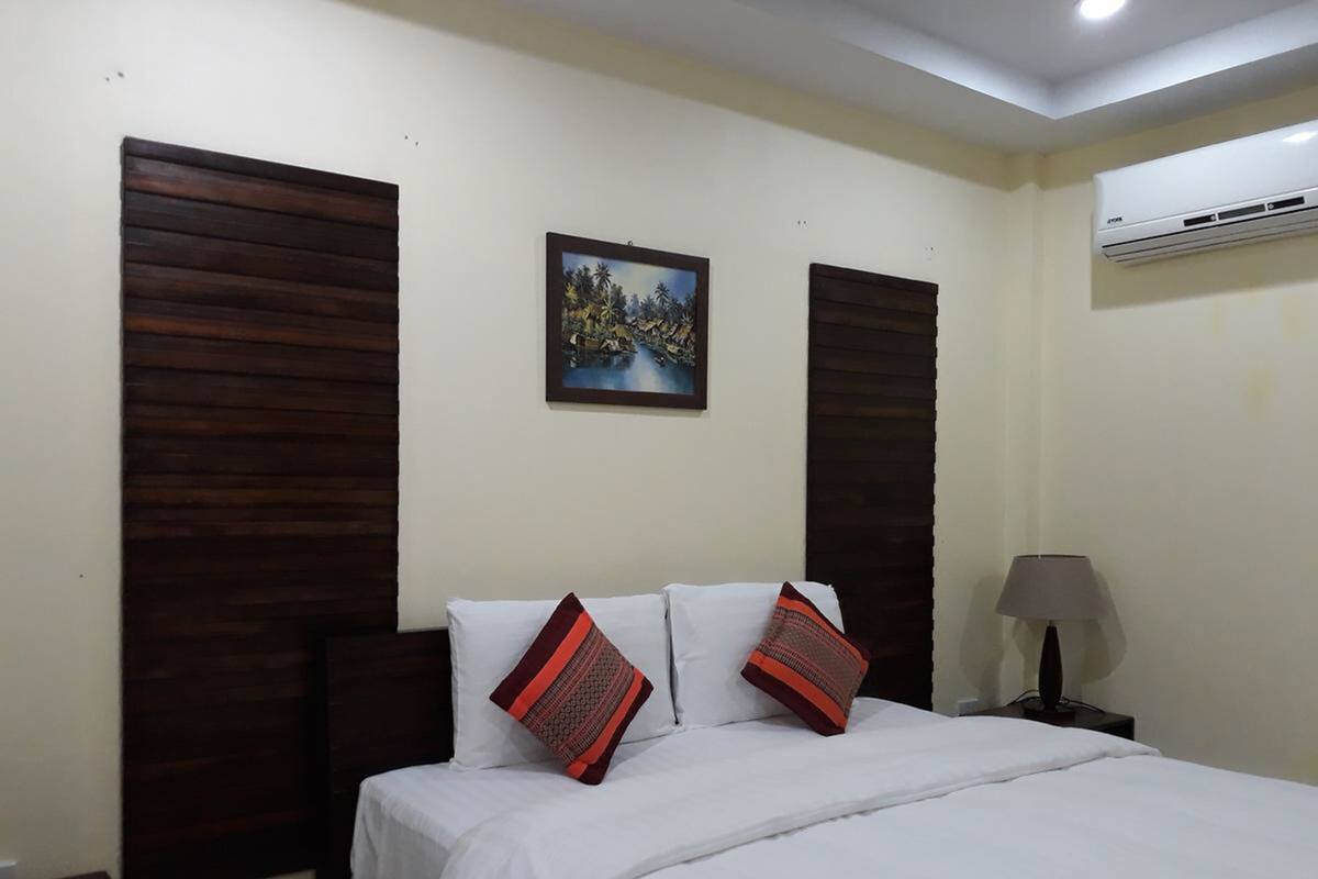 A hotel room with a bed, two pillows, a nightstand, a lamp, and an air conditioning unit.