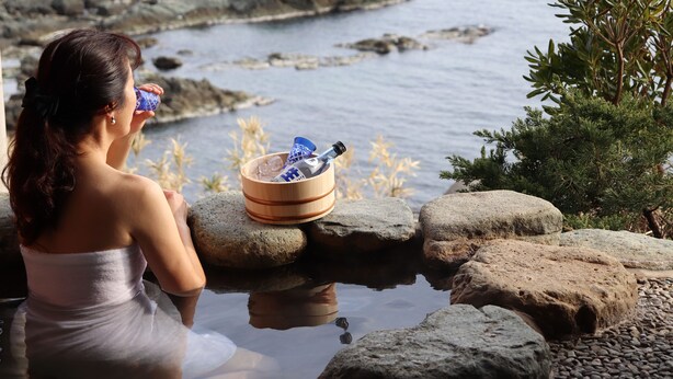 Une personne est assise au bord d’un plan d’eau, entourée de rochers et de verdure, et tient une boisson et un sac.