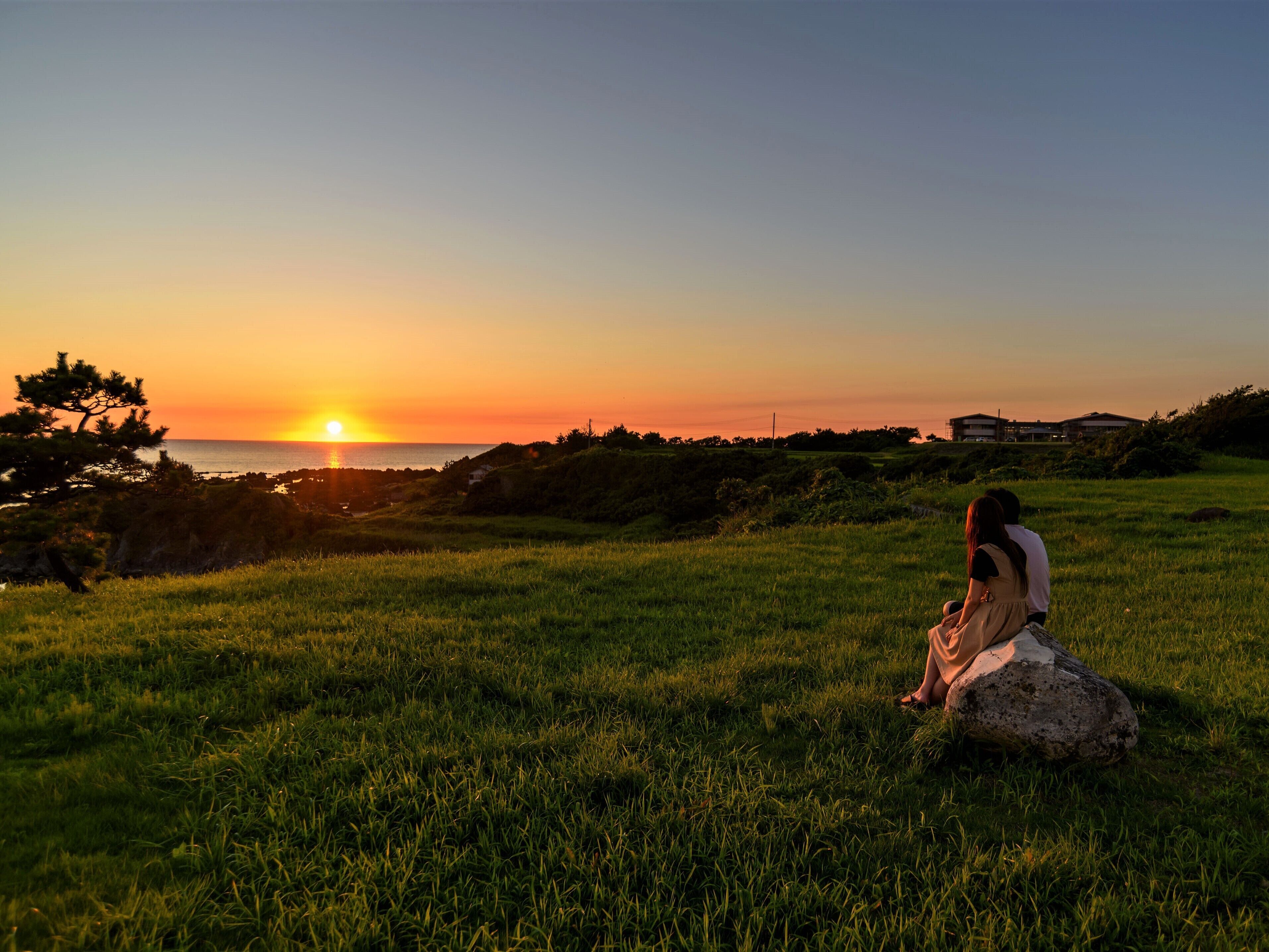 A sunset over a grassy field with a person sitting on a rock.