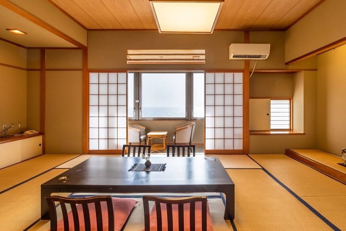 A traditional Japanese room with tatami flooring, a low table with chairs, sliding doors, and a view through a window.