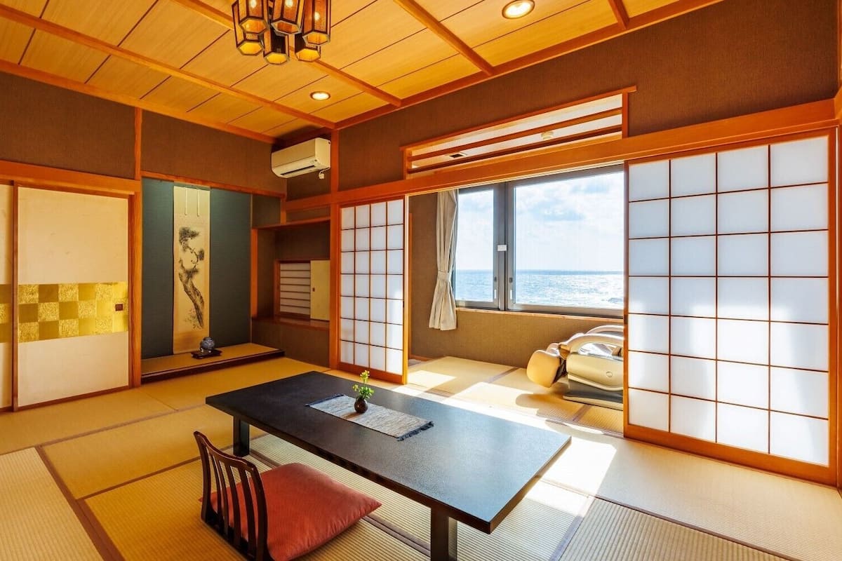 A traditional Japanese room with tatami flooring, shoji screens, a tatami mat table, a low wooden table, and a view of the sea.