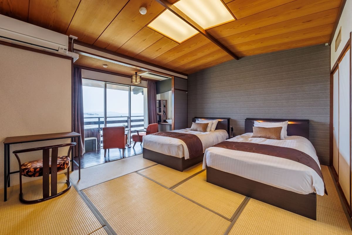 A hotel room with two beds, a small table, a chair, and a view of the sea.