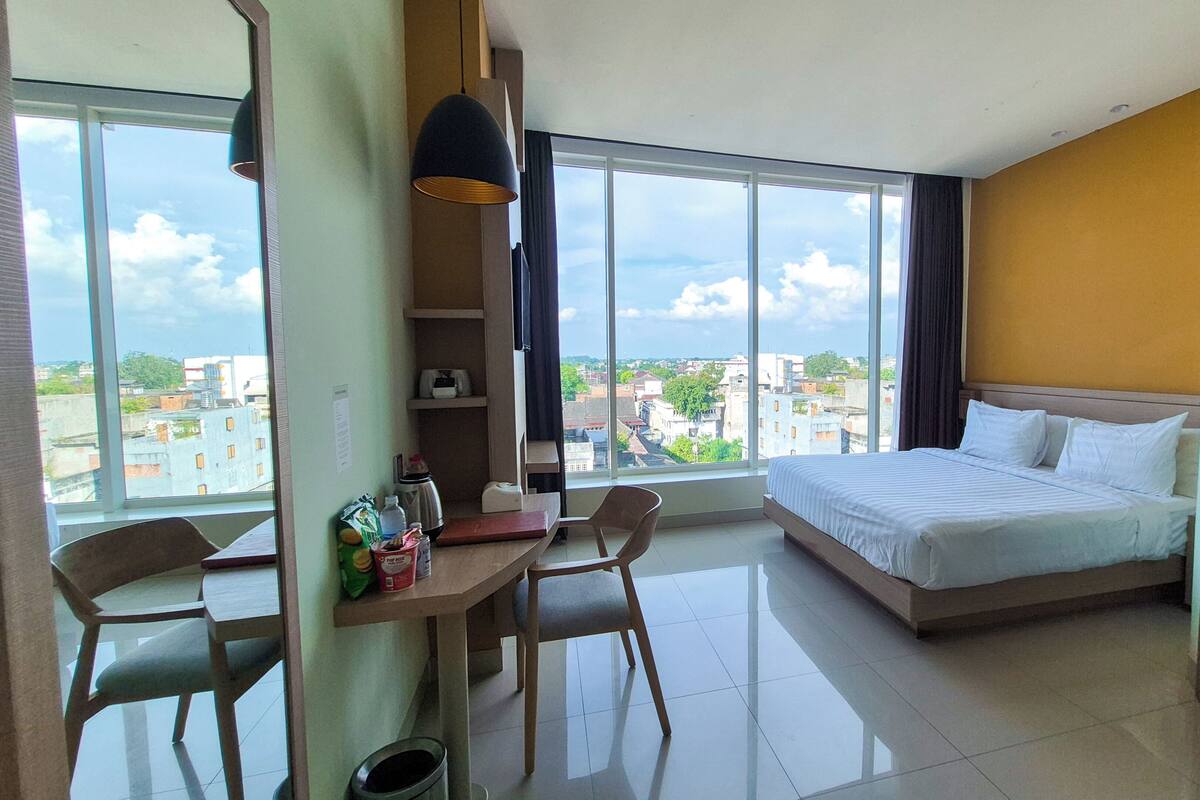 A hotel room with a bed, a desk, a chair, a large window, and a view of the city.