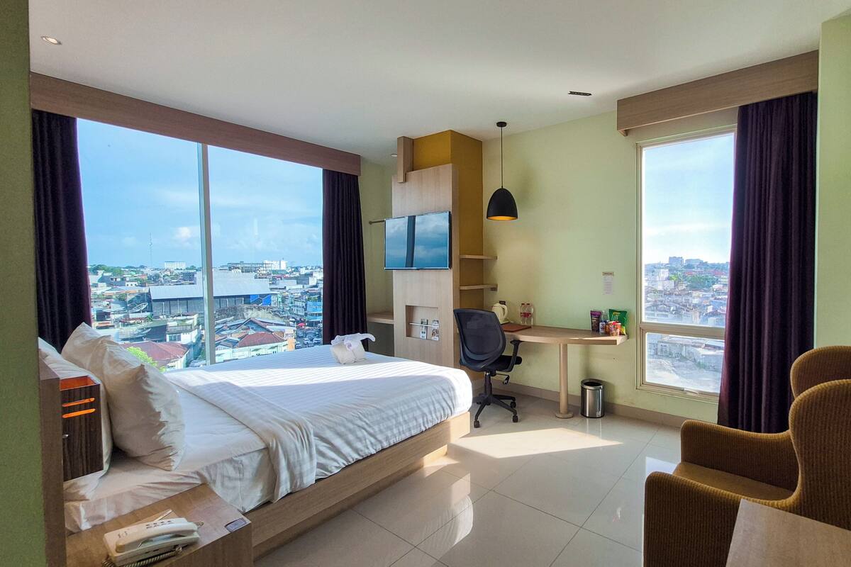 A hotel room with a bed, desk, chair, and large windows offering a city view.