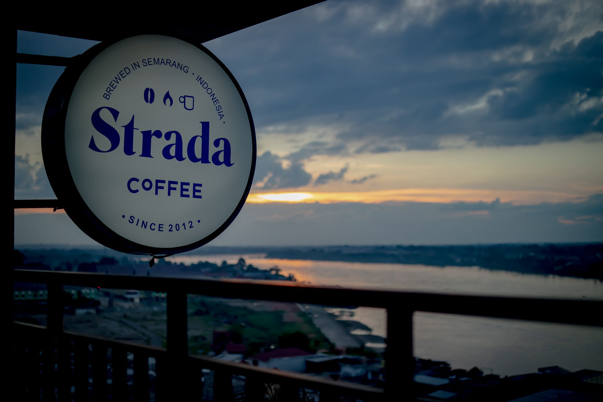 Une enseigne pour Strada Coffee, avec une vue panoramique sur un coucher de soleil se reflétant sur une étendue d’eau.