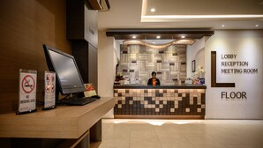 Ein Hotel-Rezeptionsbereich mit einem Holztisch, einem Computer und einer Rezeptionistin. Schilder weisen auf ein Rauchverbot hin und zeigen eine Etage an. Der Empfangstresen hat ein Fliesenmuster und eine Beschilderung für Lobby, Rezeption und Konferenzraum.