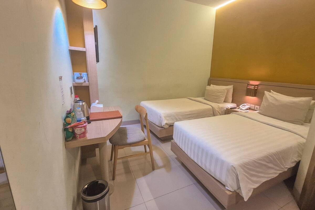 A hotel room with two beds, a desk with a chair, a trash bin, and a small table with various items.