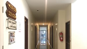 Un largo pasillo bien iluminado con puertas a ambos lados que dan a habitaciones. Hay letreros en las paredes que indican los números de las habitaciones y un extintor colgado en la pared.
