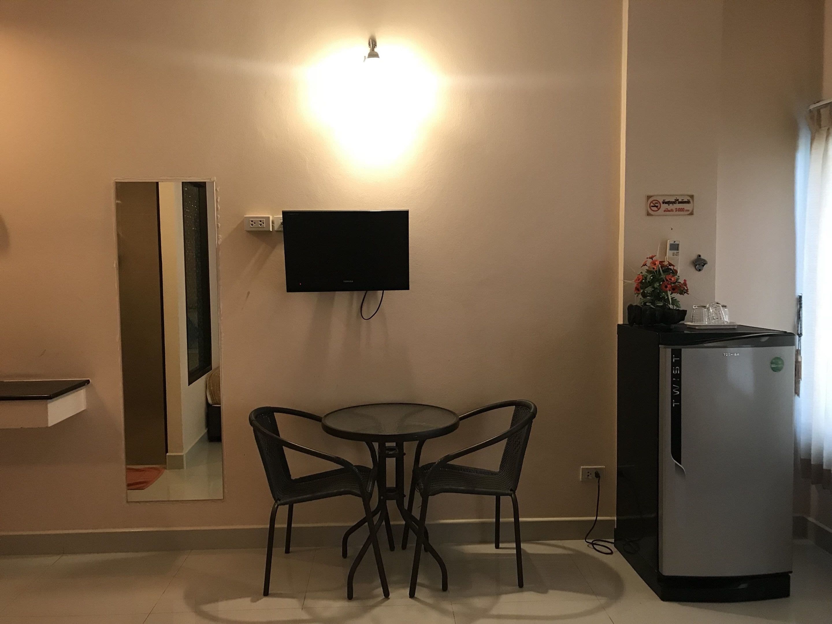 Una habitación compacta con televisión de pantalla plana en la pared, una pequeña mesa de comedor con dos sillas, un refrigerador y un espejo.