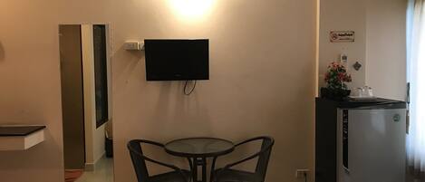 Una habitación compacta con televisión de pantalla plana en la pared, una pequeña mesa de comedor con dos sillas, un refrigerador y un espejo.