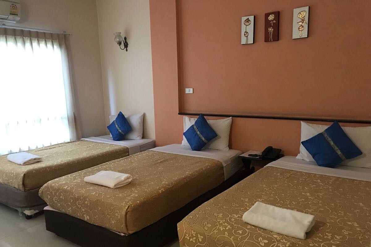 Une chambre d’hôtel avec trois lits, chacun recouvert d’une couvre-lit à motifs et agrémenté d’oreillers bleus. Un climatiseur mural est fixé au-dessus de la fenêtre, et une applique murale éclaire la pièce.