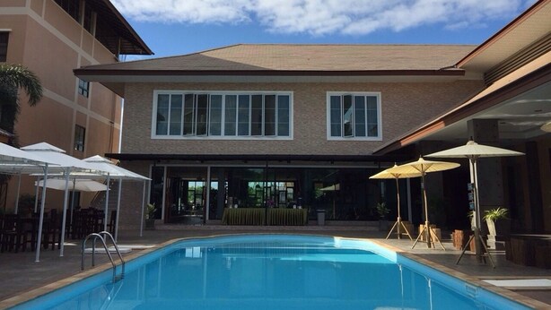 Una piscina con agua azul cristalina, rodeada de sombrillas y sillas, y un edificio con ventanas grandes y techo plano al fondo.