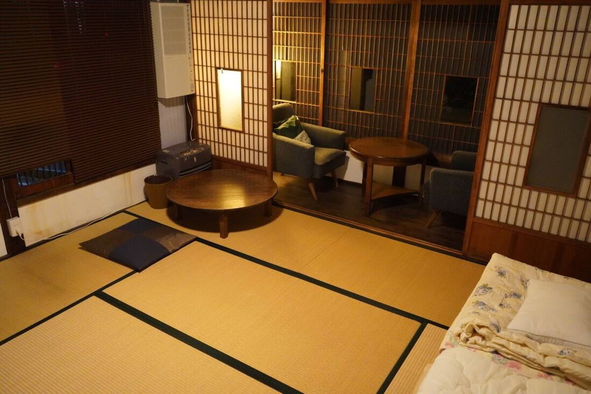 A tatami mat room with a tatami mat floor, a round table, a sofa, and a bed with a floral bedspread.