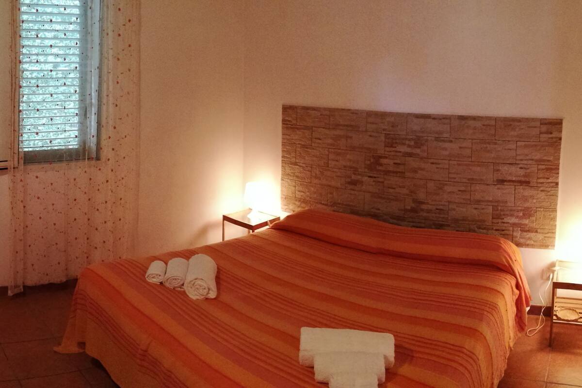 A bedroom with a bed, a wooden headboard, a nightstand, a lamp, and a window with blinds.