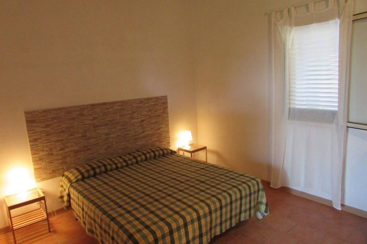 A bedroom with a bed, a nightstand, a lamp, a window with curtains, and a brick-patterned wall.