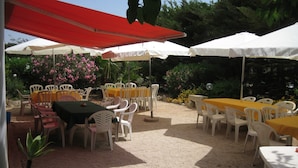 An outdoor patio with tables and chairs under umbrellas, surrounded by greenery and flowers. - L'Araucaria (Santa Croce Camerina)