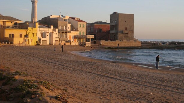 A beach with buildings in the background, including a lighthouse. - L'Araucaria (Santa Croce Camerina)