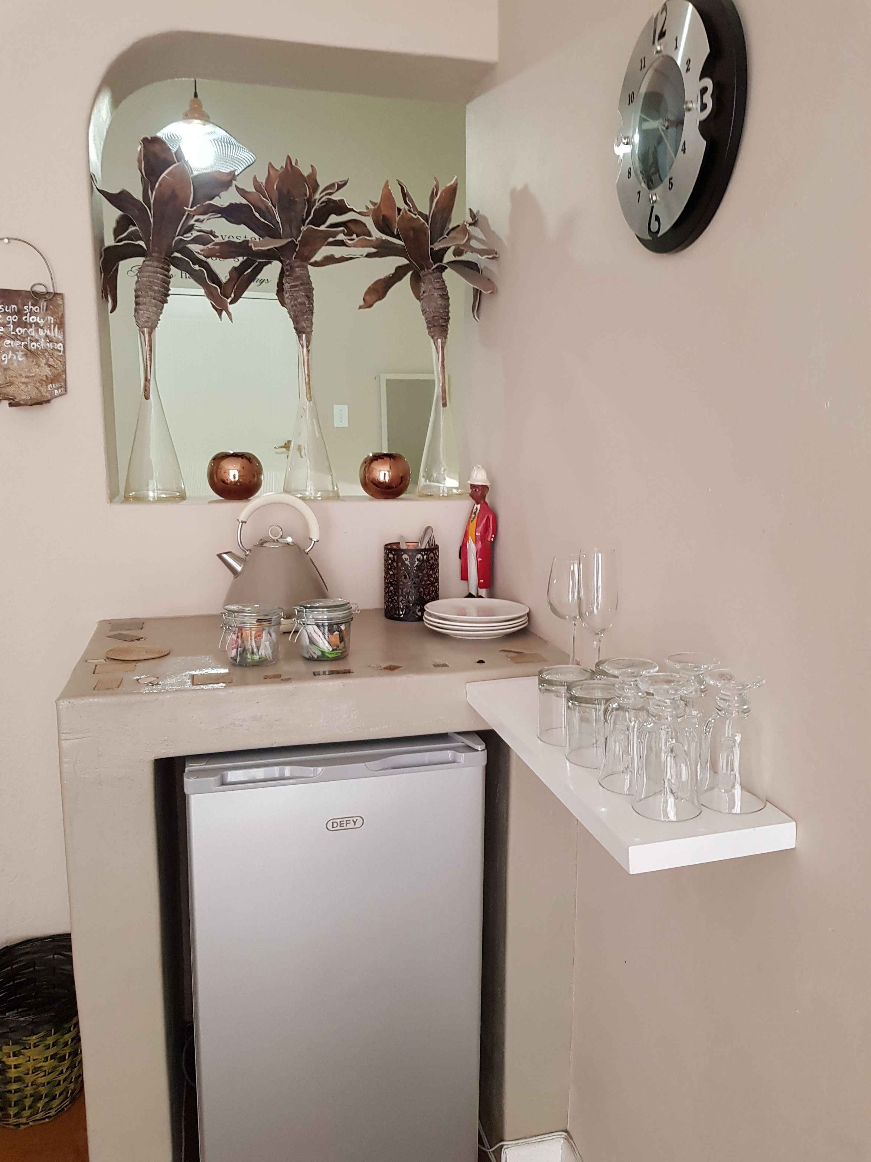 A kitchenette with a refrigerator, a sink, and a mirror above it. There are various kitchen items on the counter, including a kettle, a jar, and a decorative figure.